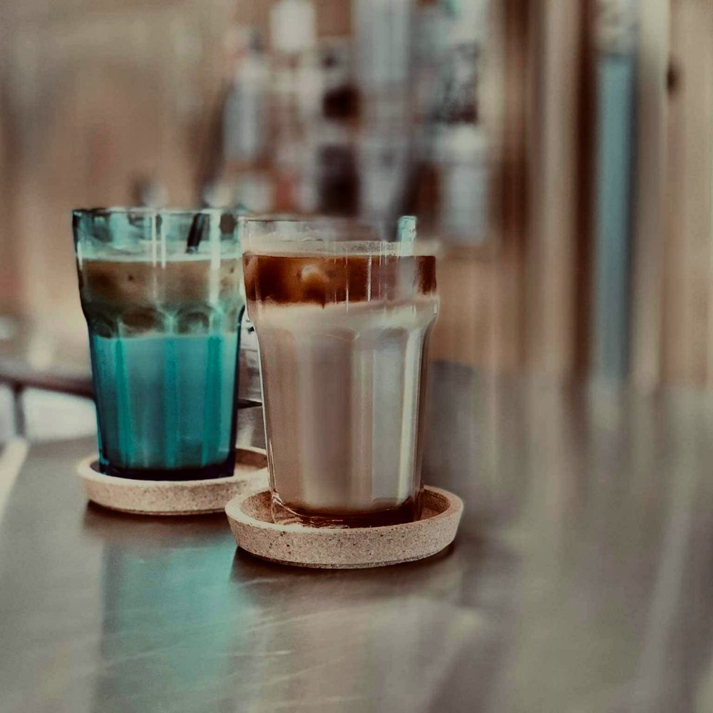 Two iced coffees in tall glasses on cork coasters, one in a turquoise glass